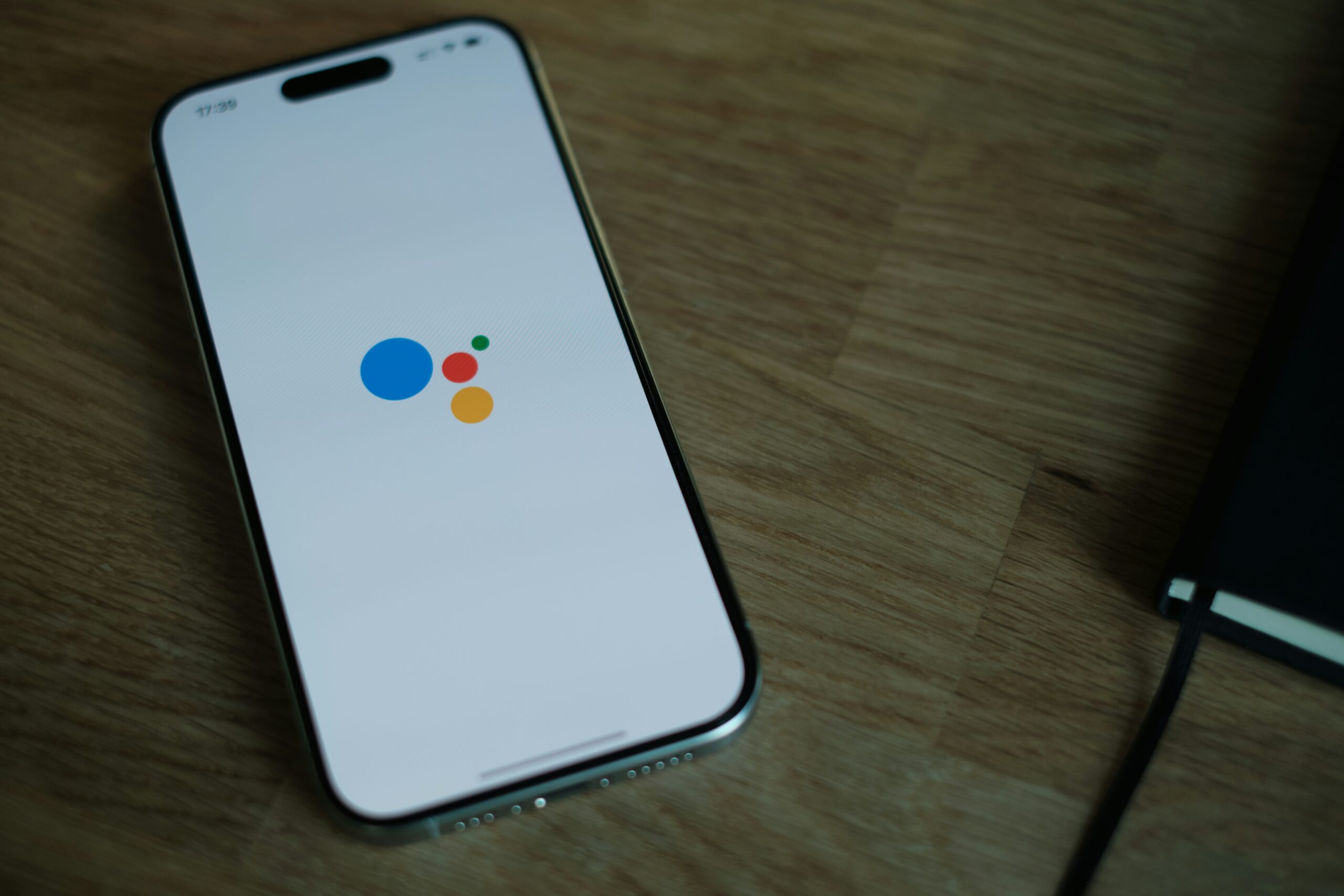 A smartphone displaying the Google Assistant interface with colored dots, symbolizing voice search technology, placed on a wooden desk next to a notebook.