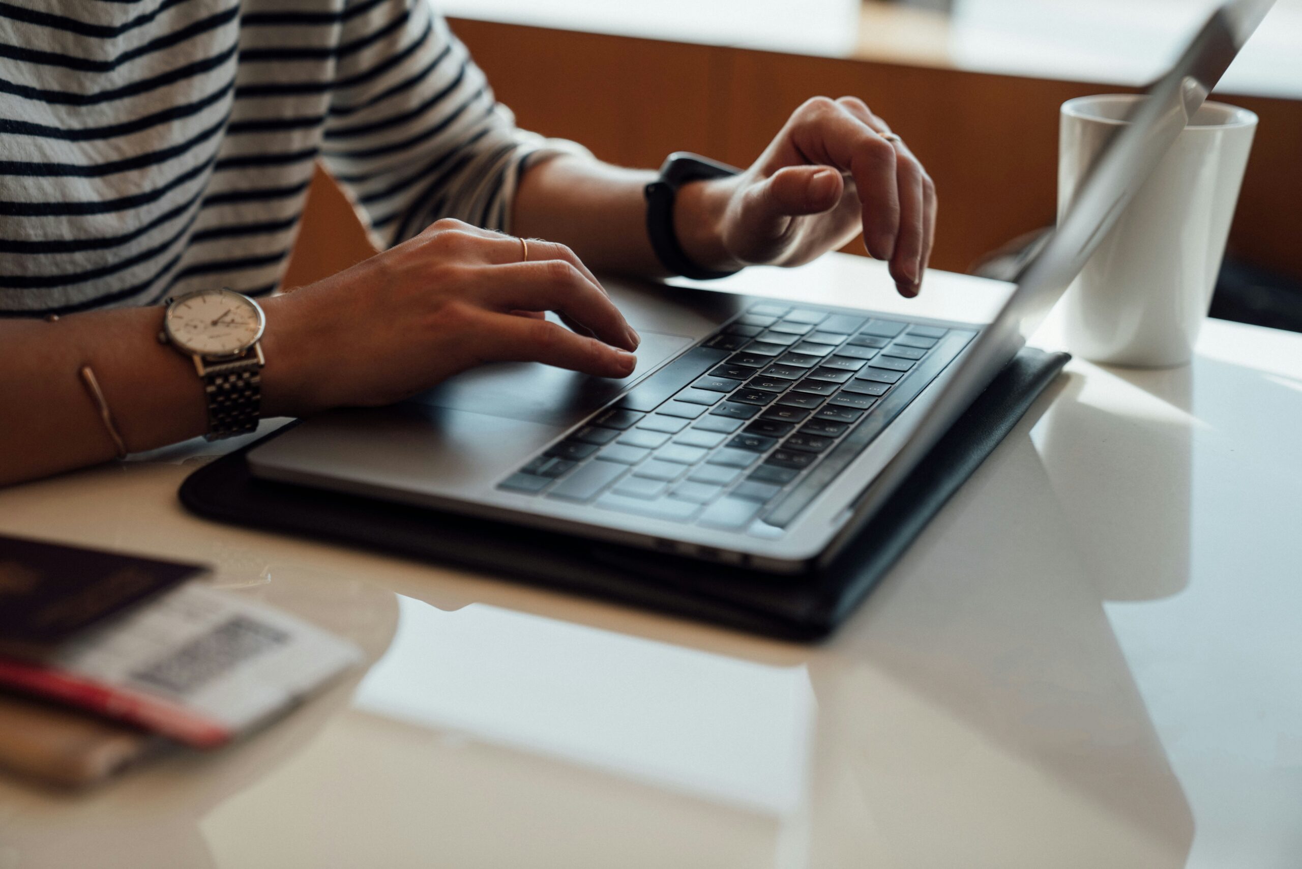 A person typing on a laptop with a watch and bracelet on his wrist, a coffee mug, and a passport nearby on a white table, symbolizing the productivity and technical work associated with dark pool indexing and technical SEO.