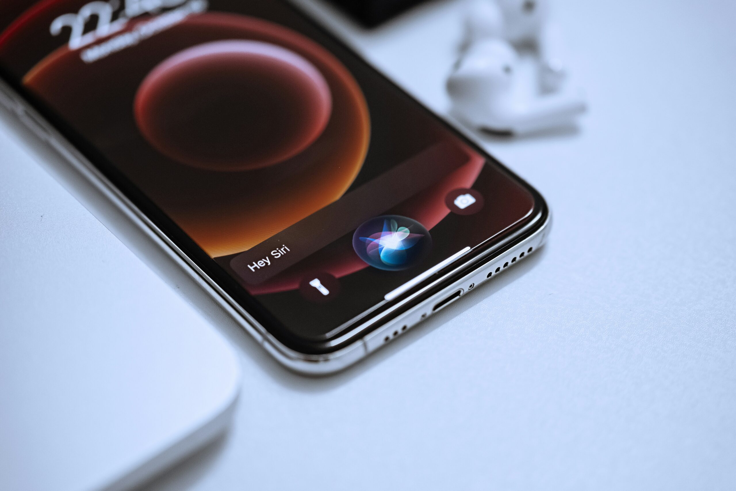 A smartphone displaying the Siri voice assistant interface with the "Hey Siri" prompt is placed on a desk next to earbuds and a laptop, highlighting voice search technology.