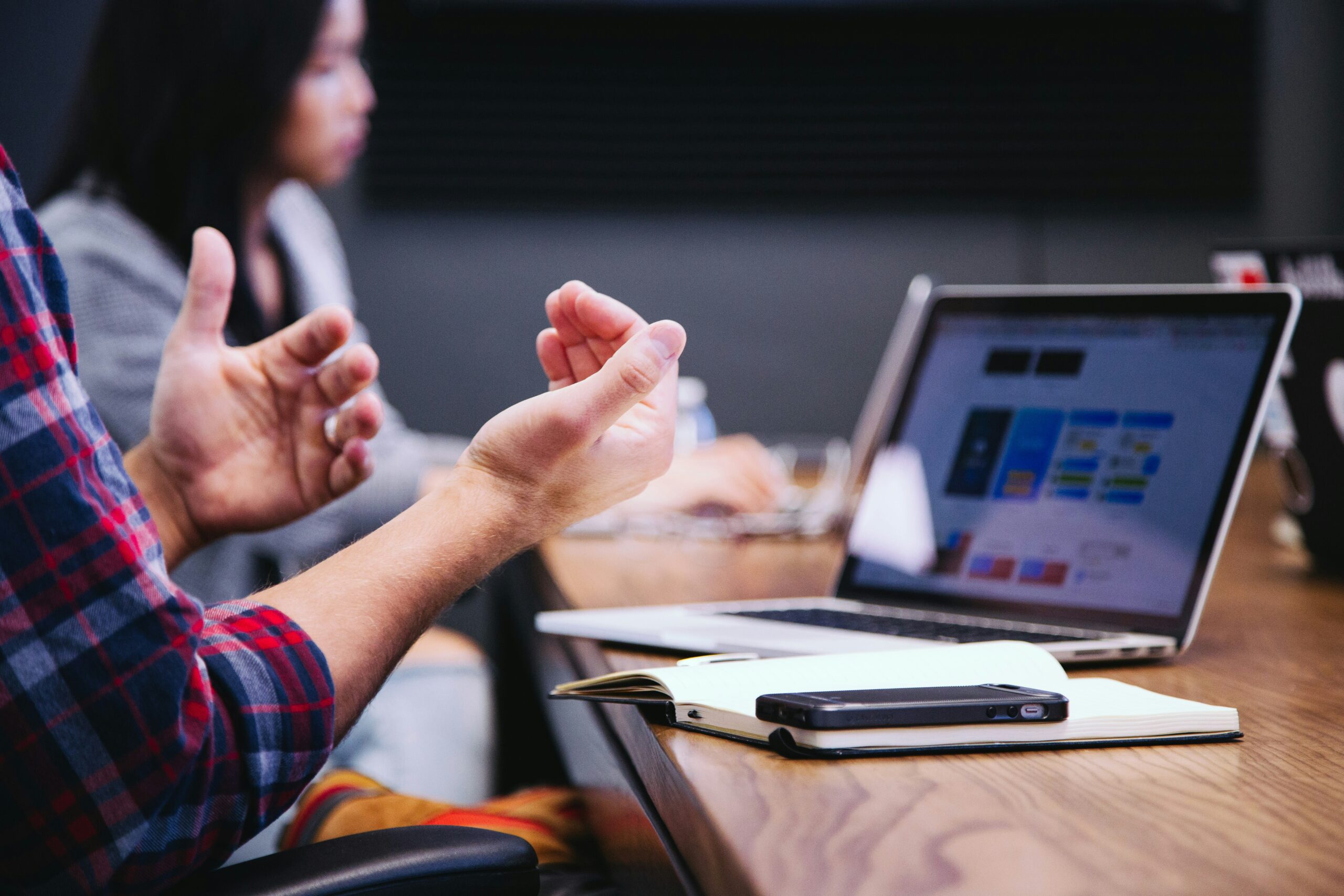 A team meeting with a person gesturing during a discussion, a laptop displaying a webpage, and a notebook with a smartphone on a wooden table.